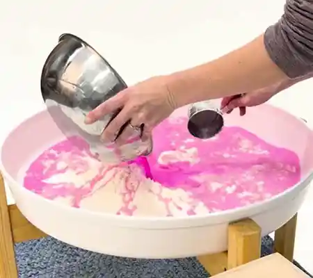 ​​Pouring water dyed with Becker&rsquo;s Washable Liquid Watercolor over cornstarch in Tuff Tray​