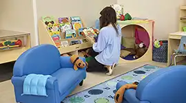 Woman in preschool classroom kneeling at bookshelf, looking at book.