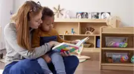​​Preschool teacher holding toddler while reading board book​